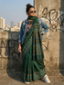 Woman in a green saree with a denim jacket standing against a graffiti-covered wall with cityscape in the background.