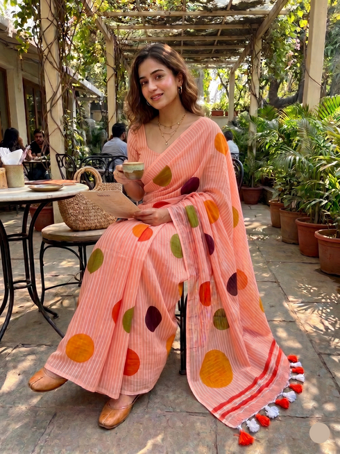 Woman in a pink saree with colorful polka dots sitting outdoors.