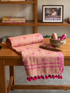 Pink and red patterned fabric with tassels on a wooden table, with books and a photo frame in the background.