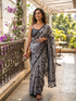 Woman in a gray saree with black polka dots on a balcony with plants and flowers.