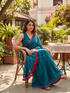 Woman in a blue saree with red border sitting outdoors in a garden setting.